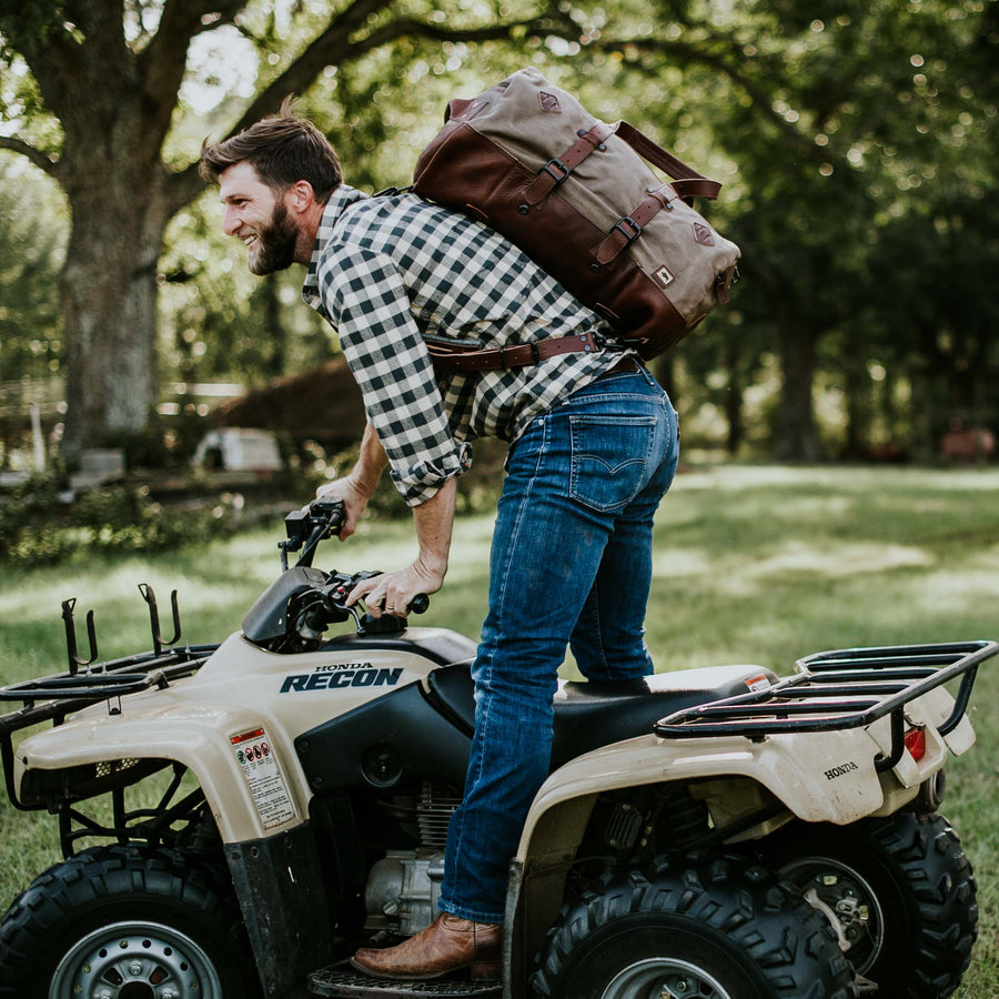 A waxed canvas duffle bag with chestnut brown leather accents, conversion straps, and a field khaki color, displayed on a wooden surface with a mountainous backdrop.